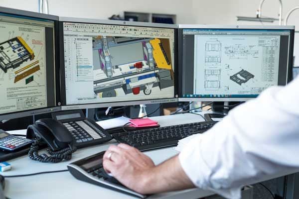 An engineer views a complex technical drawing and parts list of a construction on three monitors. 