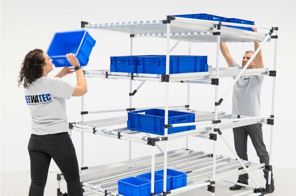 Two people store and retrieve blue KLT boxes at a BeeWaTec flow rack - for an optimized material flow